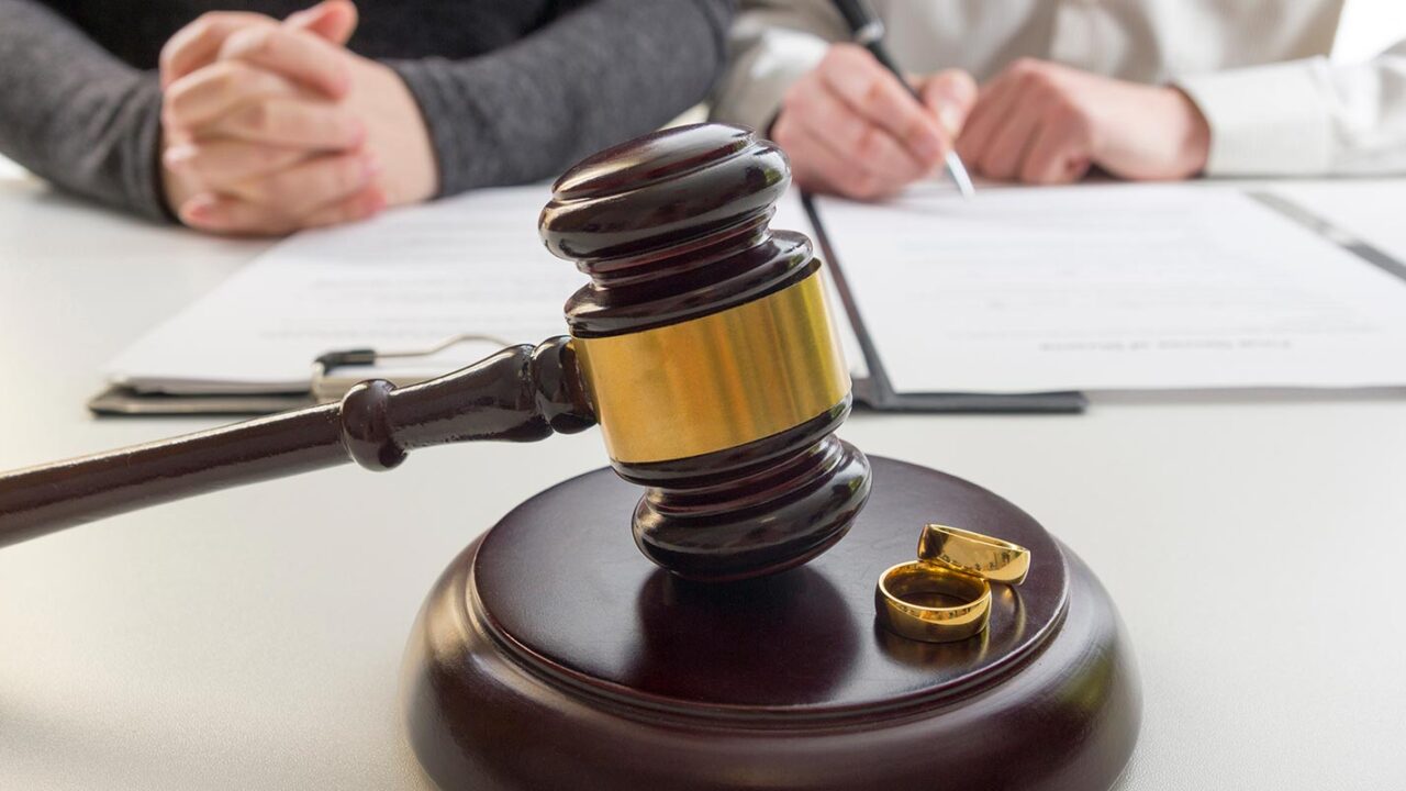 wedding rings next to a judge gavel and a couple in the background signing divorce paperwork