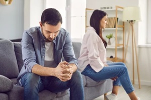 upset young man sitting on the sofa and a woman in background ignoring each other at home