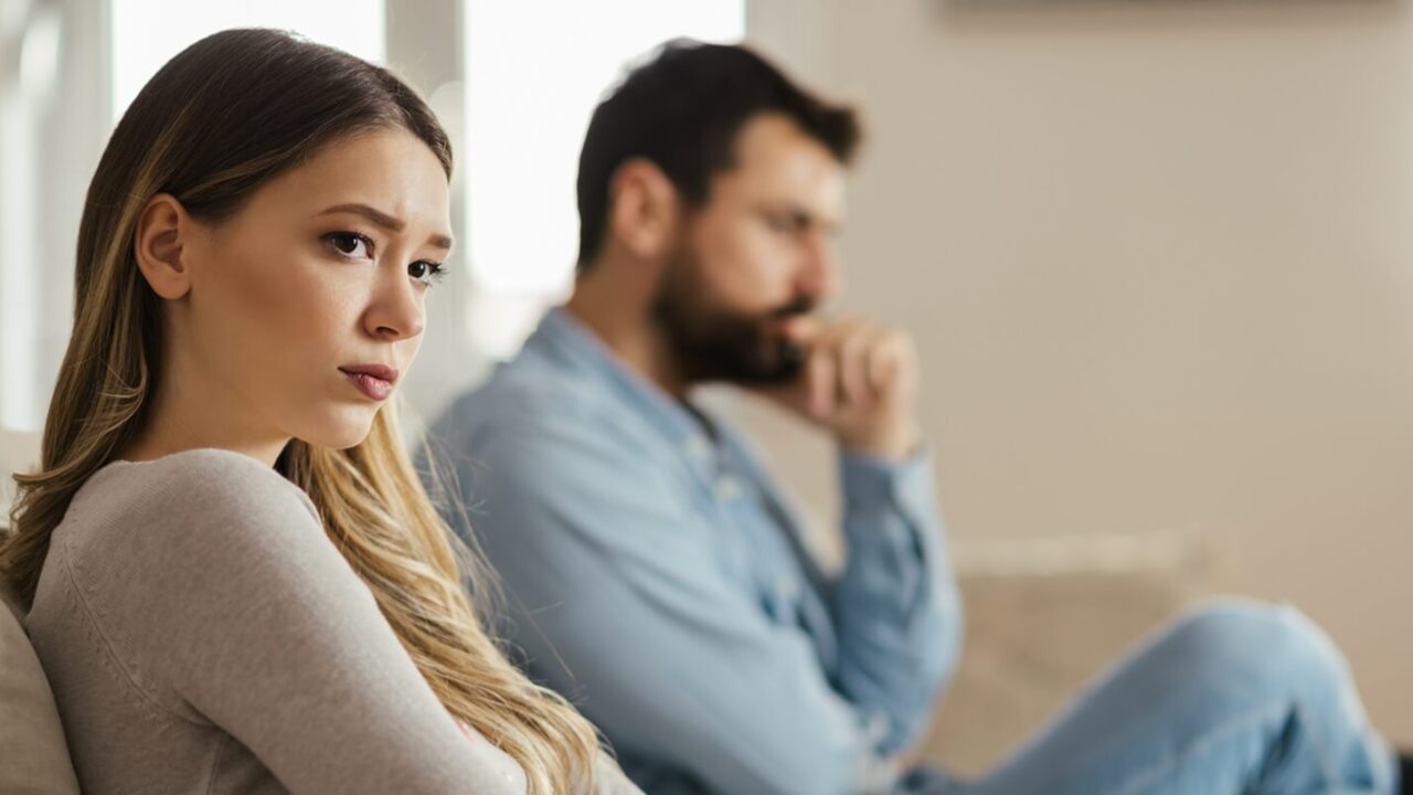 worried young woman sitting on sofa at home and ignoring her boyfriend who is sitting next to her