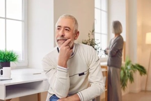 upset senior old man sitting on chair with a woman wife in background ignoring each other at home