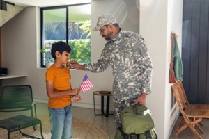 father and son leaning forward and kissing son's cheek in foyer with duffle bag and american flag