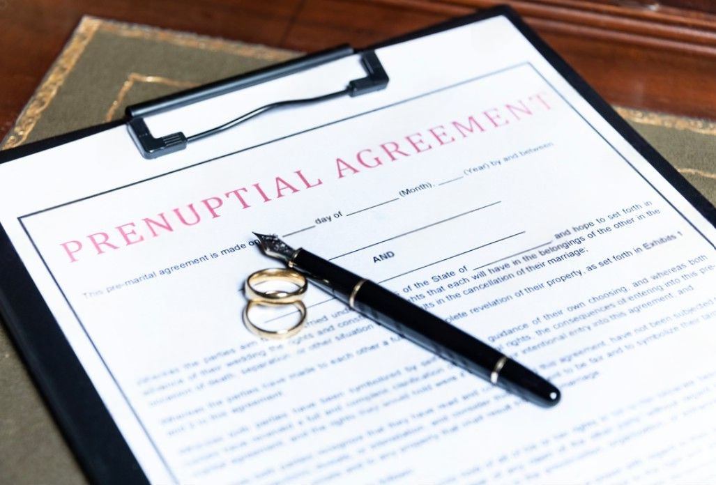 Prenuptial agreement document on a clipboard with two gold wedding rings and a fountain pen on a wooden desk
